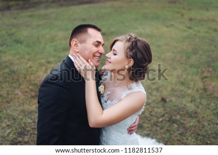 Beautiful and stylish newlyweds are hugging against the background of a green field and forest. A wedding portrait of an adult groom in a black suit and a cute bride in a lavish dress. Wedding.