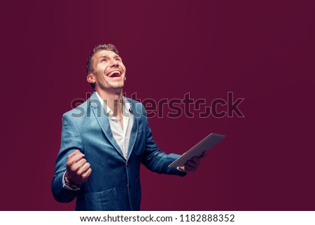 Portrait of Successful Young Stilish Man in Suit holding tablet with Fist up. Isolate. Red background. Winner.
