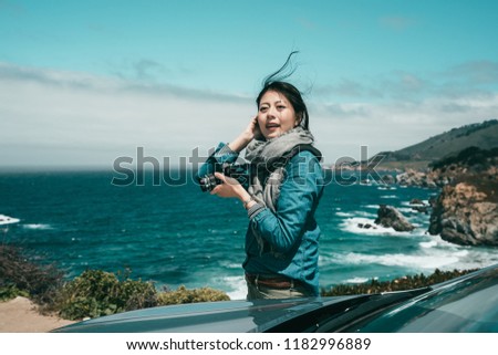a beautiful woman taking photos of the spectacular ocean and cliff standing with her car nearby.