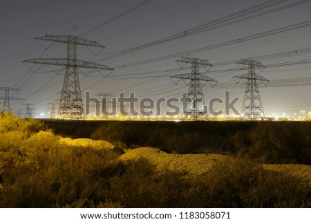 High voltage electric pylon towers & power cable lines at night with city light as background installed at a desert