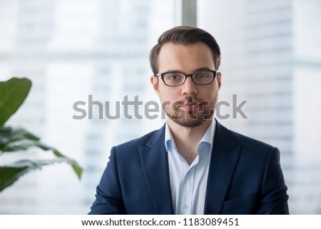 Portrait of serious millennial businessman wearing glasses looking at camera, headshot of concentrated confident male worker or director posing in modern office, making photo or picture near window