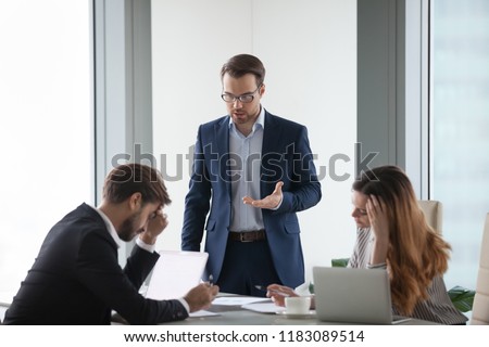 Angry male worker standing talking to colleagues lecturing on mistake in documents or failed project, businessman show dissatisfaction to business partner during negotiations or meeting in office Royalty-Free Stock Photo #1183089514