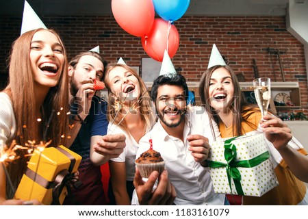 Group of friends holding gifts, sparklers and raising glasses with champagne, multicultural bearded man holding birthday cake, they are celebrating birthday