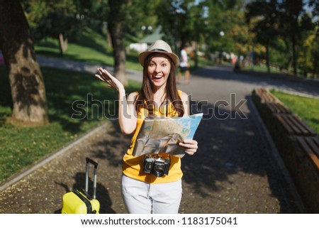 Young smiling traveler tourist woman in yellow clothes, hat with suitcase spread hands holding city map in city outdoor. Girl traveling abroad to travel on weekends getaway. Tourism journey lifestyle