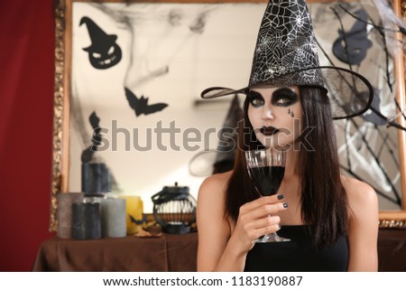 Beautiful woman dressed as witch with wine in room decorated for Halloween