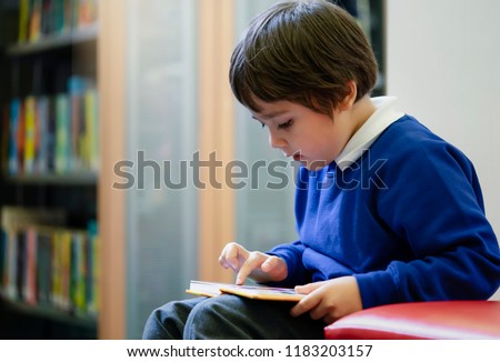 Preschool kid sitting on red sofa enjoy reading a cartoon in the library in the school,Cute boy with excited face while reading favourite  book in library.Early year student activity concept