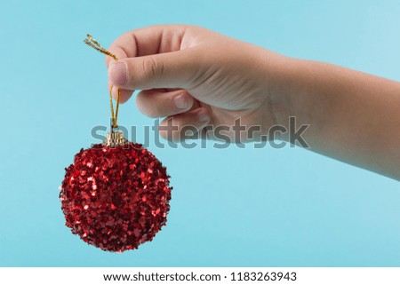the child's hand holds a red decorative ball, a Christmas tree toy, on a pastel turquoise background, a minimal concept