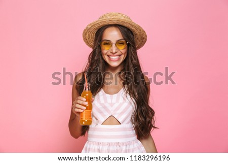 Photo of happy woman 20s wearing sunglasses and straw hat drinking juice from glass bottle isolated over pink background