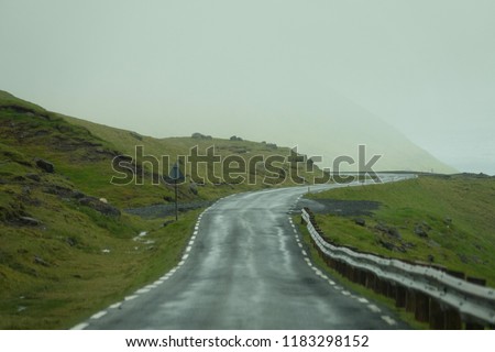 Empty concrete road winds along the steep grassy hill of a mountain covered in thick fog. Stunning shot of dense white mist gathered over the scenic road in the rugged countryside of Faroe Islands.
