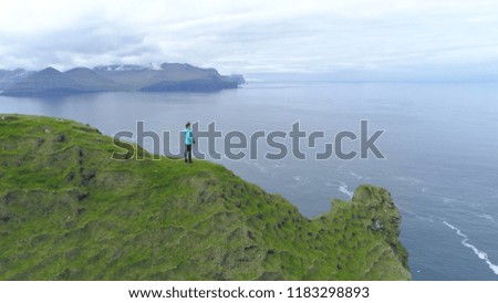 AERIAL, COPY SPACE: Flying over beautiful Scandinavian landscape and unrecognizable female photographer observing the vast sea. Spectacular aerial shot of the rugged seaside in scenic Faroe Islands.