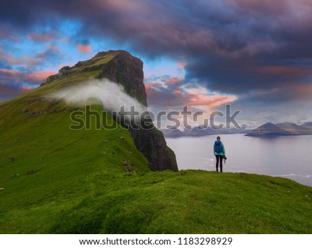 Female photographer stands on the edge of a cliff and watches the spectacular blue and orange evening sky. Stunning view of the magnificent green cliffs and mountains in the picturesque Faroe Islands.