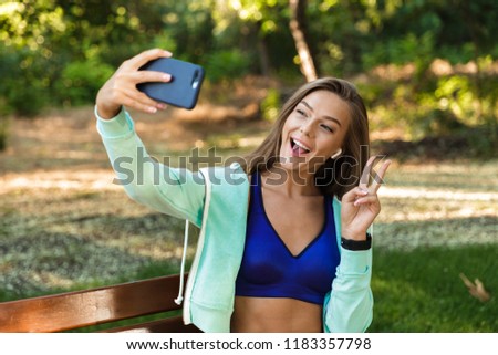 Photo of pretty young sports woman in park outdoors listening music with earphones take a selfie by mobile phone make peace gesture.