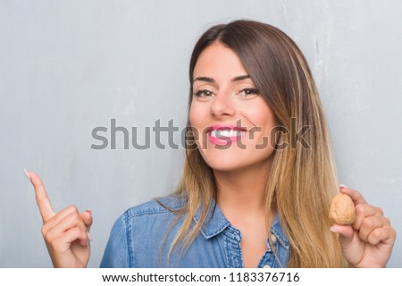 Young adult woman over grey grunge wall showing dried walnut very happy pointing with hand and finger to the side