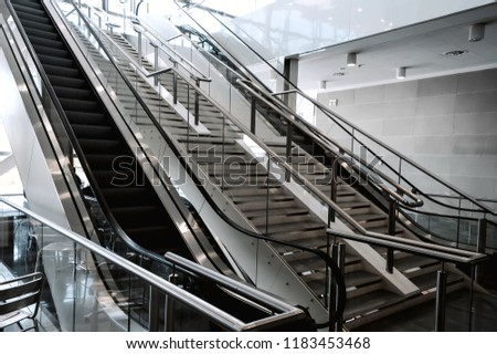 Escalators in big modern area. No people around. Grey tones interior design with shiny glass. Perspective side view. Natural light illumination. Mall navigation and connection.