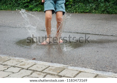 This image shows a healthy young boy splashing in the puddle after a warm summer rain. The boy is in blue shorts, the water spouts high away. You can use it to express happiness, heath and joy of life