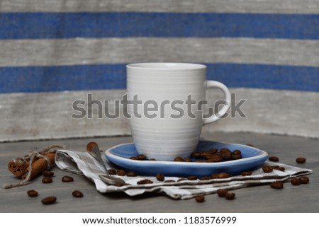 A cup of coffee on a saucer with coffee beans & a spoon nearby on a checkered napkin, two cinnamon sticks nearby & coffee beans around on a wooden surface & on a background of a striped linen fabric