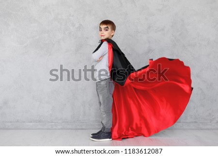 Boy profile portrait wearing Halloween vampire makeup and fluttering his red, black cloak, on a gray textured background. Cute kid wearing super hero costume with crossed hands, copy space available.