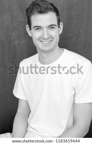 Model headshot of a man in a t-shirt against a textured black background looking at the camera and smiling. Clean cut white man posing for the camera on black background, black and white.