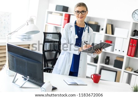 A young girl in a white coat is standing near a table in her office and holding a tablet and a pen. A stethoscope hangs around her neck.