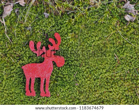 Red wooden silhouette of moose, or elk, or deer, lies on lush green forest, swamp moss with dry grass on top edge of picture; with copy space