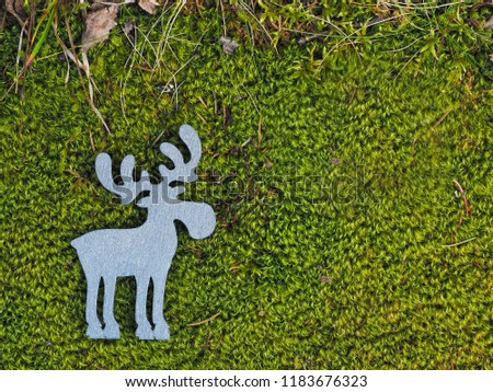 Silvery wooden silhouette of moose, or elk, or deer, lies on lush green forest, swamp moss with dry grass on top edge of picture; with copy space