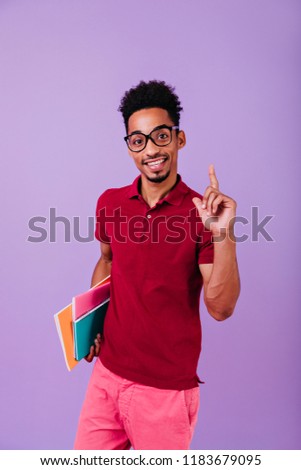 African student in red attire posing with interested smile. Studio photo of good-humoured black man in glasses holding books and expressing happiness.