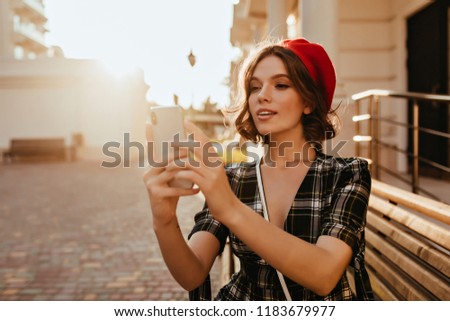 Cute dark-haired lady taking picture of herself in good sunny day. Portrait of jocund female model in stylish french beret holding smartphone.
