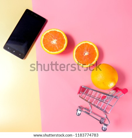 Square picture flat lay black mobile phone on top left two pieces of slice orange whole orange moving to small empty red trolley on bottom right on pink and orange background online shopping concept
