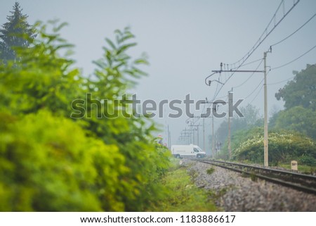 Train track with overhead catenary and electric lines in a foggy misty morning, leading into white, with a road crossing with visible van crossing the tracks
