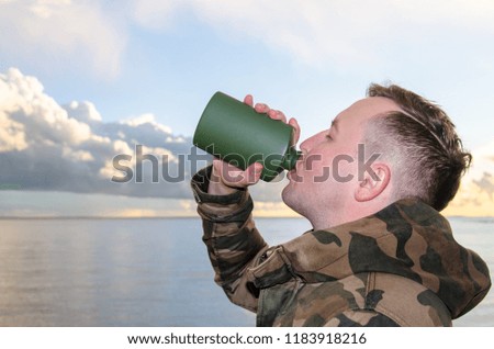 A young man in the camouflage jacket drinks water from a green jar or flask on the sea background.