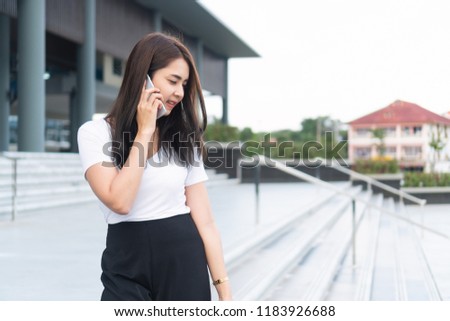 Young asian businesswoman in casual is talking on her mobile phone while walking outdoor. Beautiful and smiling female student college calling someone on phone with building background and copy space.