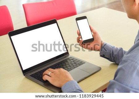 Businessman using blank white screen smartphone and empty white desktop screen laptop on wooden table at workplace. Rear view of man hand working with mobile phone and  computer. Mockup image concept.