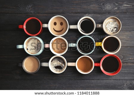Cup of delicious hot coffee with foam and smile among others on wooden background, flat lay. Happy morning, good mood, inspiration