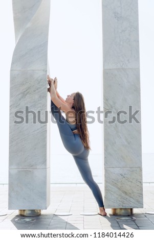 Beautiful girl with long hair dressed in sportswear doing the splits in urban exterior. Flexible woman doing yoga meditation with the white sky on the background. Calmness, relax, healthy lifestyle