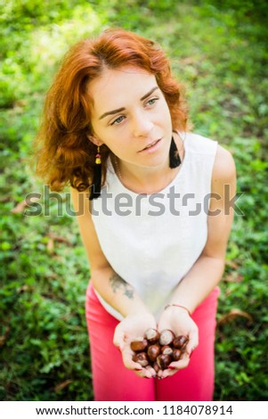 A portrait of a red curly woman holding cheastnuts on his hand. Closeup portrait. Green eyes and red hair. Green grass background.