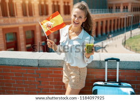 smiling modern traveller woman with trolley bag and green smoothie against Puerta De Atocha train station building