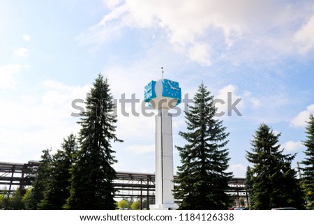 Unusual clock on a background of a white-blue sky. A clock in the form of a molecule, an atom. On chemical production, a watch-column, a clock tower in a spruce forest. Backdrop of chemical pipelines.