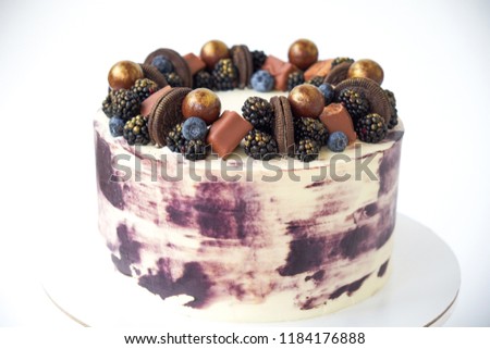 Cake with white and black cream, decorated with blueberries, blackberry, chocolate chip cookies, candies, bars and gold on a white background. Picture for a menu or a confectionery catalog.
