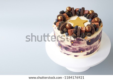 Cake with white and black cream, decorated with blueberries, blackberry, chocolate chip cookies, candies, bars and  gold star on a gray background. Picture for a menu or a confectionery catalog.