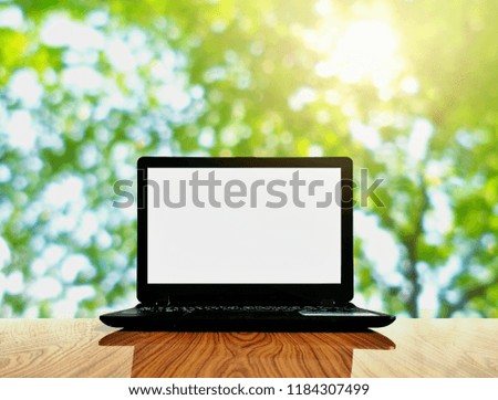 Black laptop with empty white screen on wooden glossy table and flare light with blurred background of many green leaves in the garden, technology concept with space for your text 