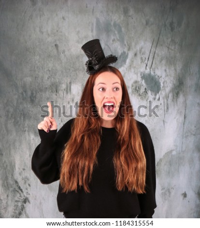 Portrait of a young woman wearing a halloween hat while pointing her finger up against a grey background