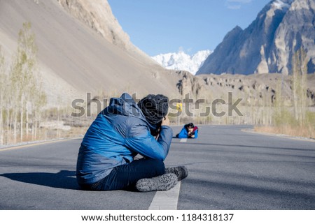 Male photographer is taking picture of other two travelers sitting on concrete road against the background of brown hills, snowy mountains, glaciers, dry forests. Concept tourism, hiking, adventure.