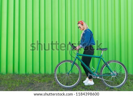 Portrait of a stylish woman in a denim jacket and sunglasses, pulling a bike on the background of a green wall. Copyspace