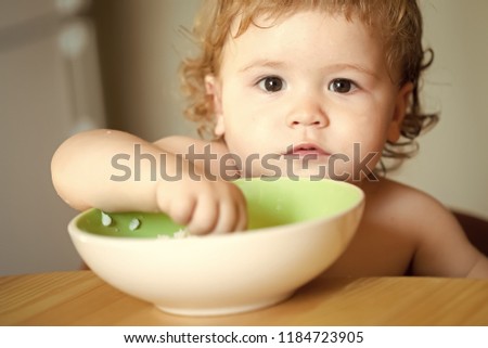 Portrait of interesting small pretty baby boy with blonde curly hair and round cheecks eating from green plate with hand closeup, horizontal picture