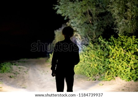 back view of man standing outdoor on night forest road lighting the way with electric flashlight