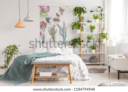 Close to nature bright bedroom interior with a bed covered with white sheets and marine blanket. Green plants on shelves next to the bed. Real photo.