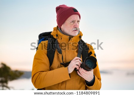 Portrait of mature man with grey beard exploring Finland in winter. Traveler Taking pictures on the top of rock. Beautiful view of northern landscape with frozen Baltic Sea and snowy islands.