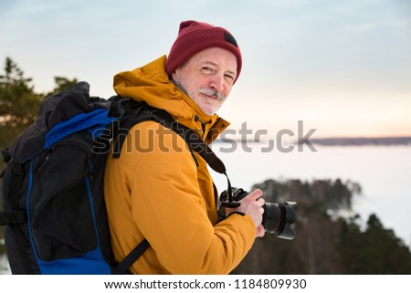 Portrait of mature man with grey beard exploring Finland in winter. Traveler Taking pictures on the top of rock. Beautiful view of northern landscape with frozen Baltic Sea and snowy islands.