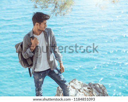 Traveler with backpack on the rocks near the sea looking away. Summer Travel Vacation. Handsome young caucasian tourist man in casual clothes outdoors on the nature.