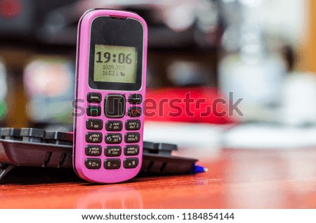 Old button mobile phone pink color on the desk near a computer keyboard. Clock and indicators on the black and white screen.